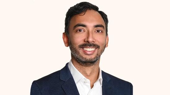A man with short dark hair and a beard smiles, wearing a navy blazer and white shirt, against a plain light background.