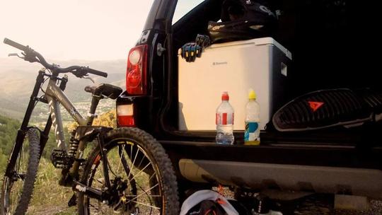 A mountain bike leaning against the open trunk of a car, with a cooler, helmet, and water bottles inside, set against a scenic mountain backdrop.