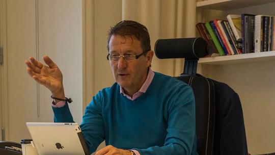 Conni Jonsson sitting at a desk, gesturing with one hand. The background features a bookshelf with various books.
