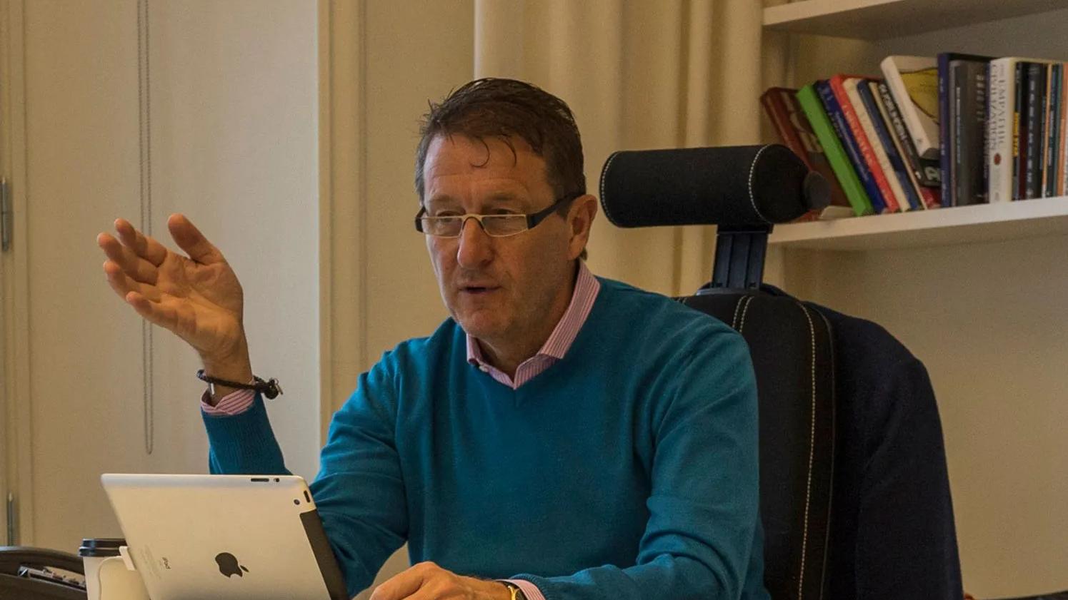 Conni Jonsson sitting at a desk, gesturing with one hand. The background features a bookshelf with various books.