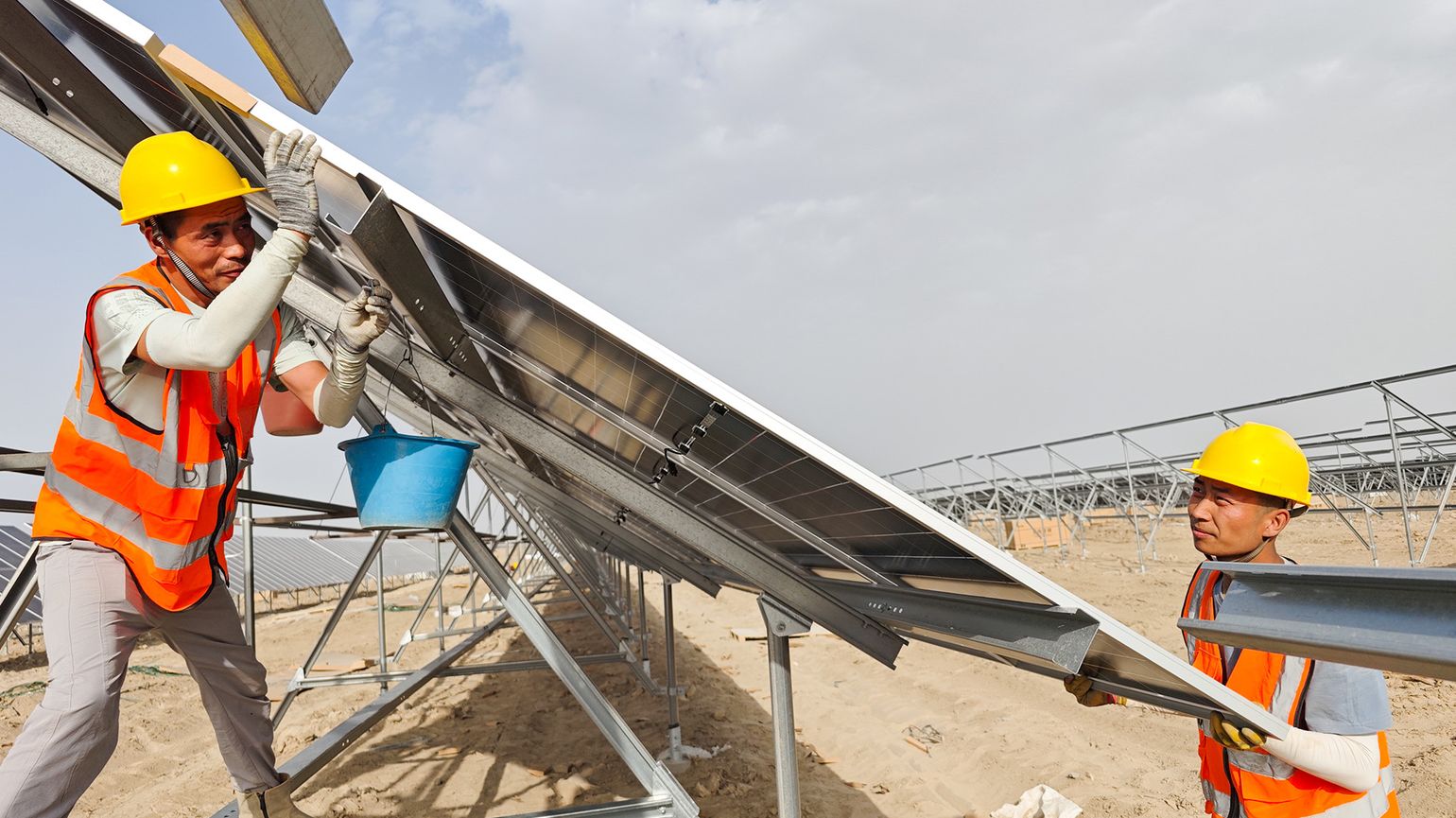 Two workers in orange safety vests and yellow helmets install a solar panel on a metal frame at a solar farm construction site.
