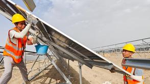 Two workers in orange safety vests and yellow helmets install a solar panel on a metal frame at a solar farm construction site.
