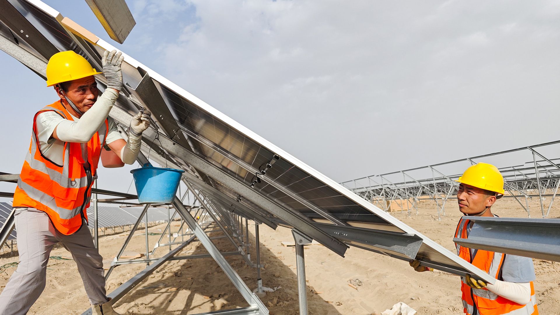 Two workers in orange safety vests and yellow helmets install a solar panel on a metal frame at a solar farm construction site.