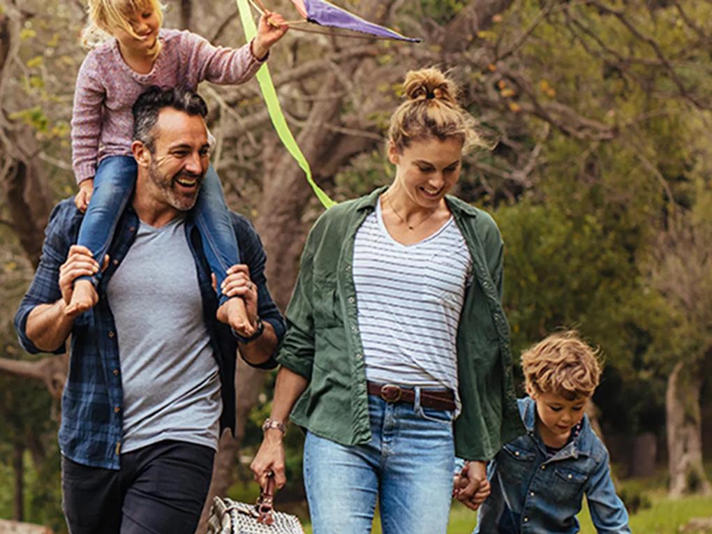 A family enjoying a walk outdoors. A man carries a young girl on his shoulders, while a woman holds hands with a boy.