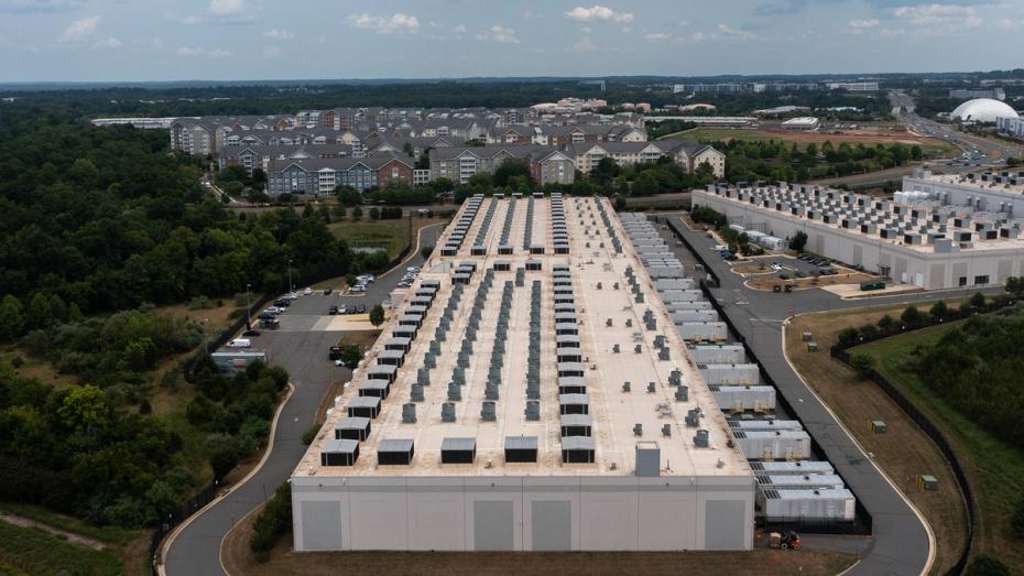 A large data center building with rows of cooling units on the roof, surrounded by trees, parking lots, and residential buildings in the background.