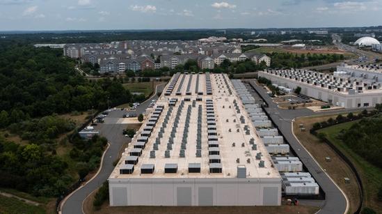 A large data center building with rows of cooling units on the roof, surrounded by trees, parking lots, and residential buildings in the background.