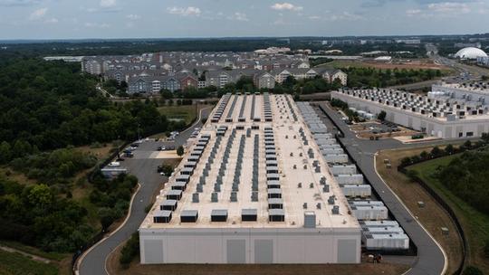 A large data center building with rows of cooling units on the roof, surrounded by trees, parking lots, and residential buildings in the background.