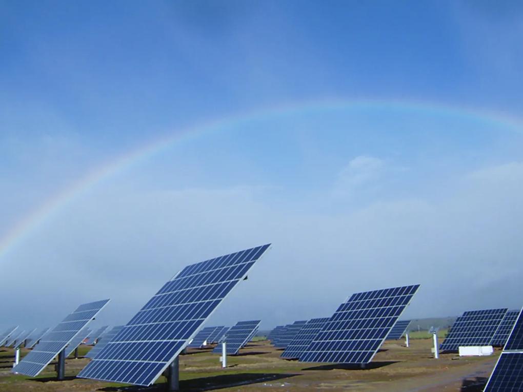 A field of solar panels under a clear blue sky with a faint rainbow arching overhead.