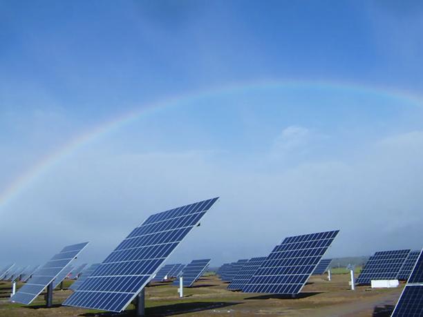 A field of solar panels under a clear blue sky with a faint rainbow arching overhead.