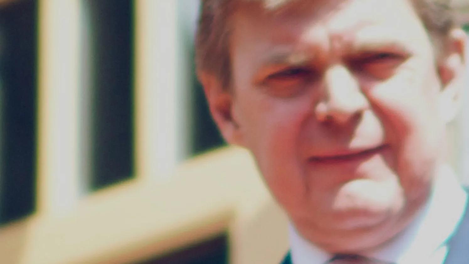 A close-up, slightly blurred image of a person in a suit, standing outdoors with a building in the background.