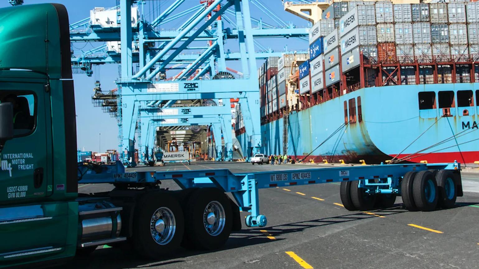 A green truck parked at a shipping port with large cranes and a container ship in the background.