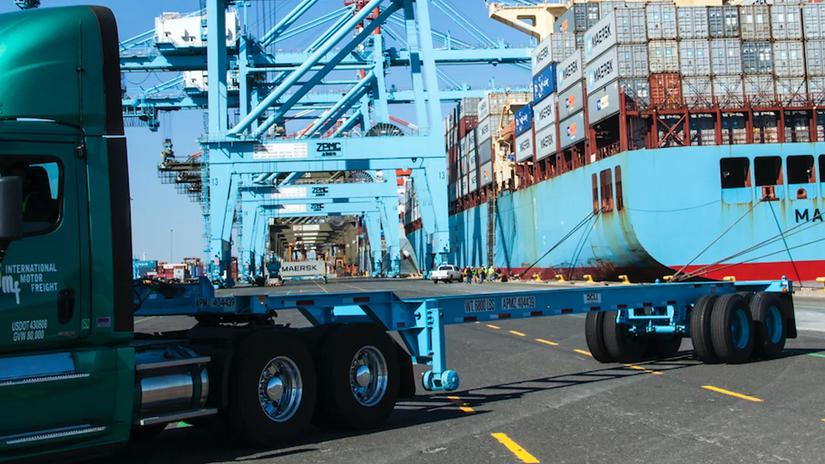 A green truck parked at a shipping port with large cranes and a container ship in the background.