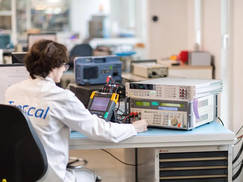 A technician in a lab coat labeled "Trescal" operates electronic testing equipment at a workstation.