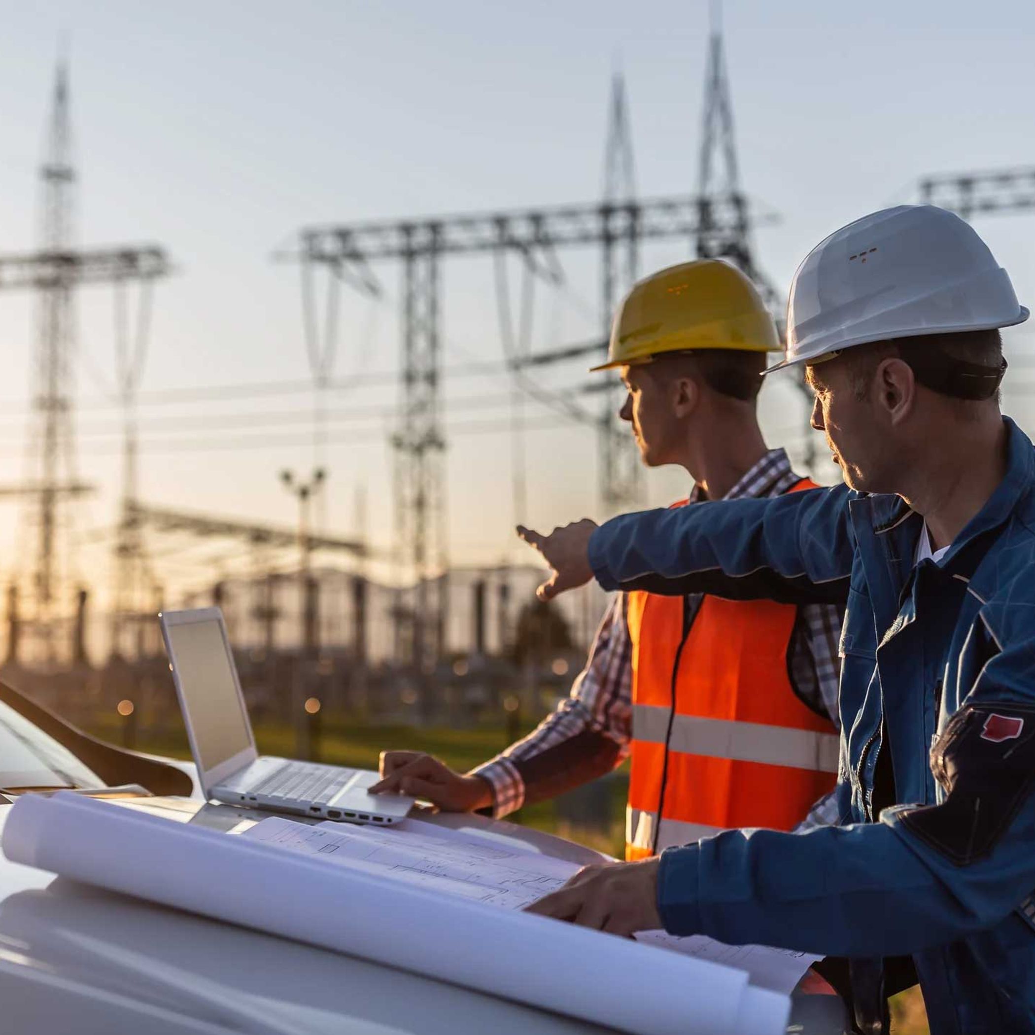 Blue collar worker points towards sun setting over power distribution center with white collar worker looking the same direction.