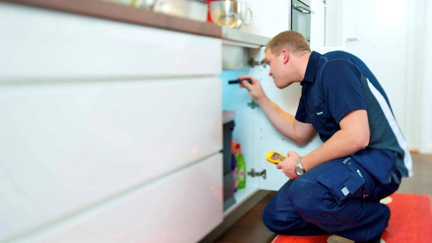 xA technician in a blue uniform inspecting under a kitchen sink, holding a flashlight and a measuring device, focused on a maintenance task.