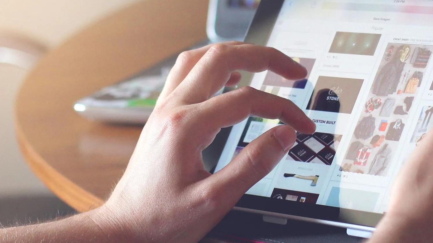 A person using a tablet, browsing through images on a wooden table.