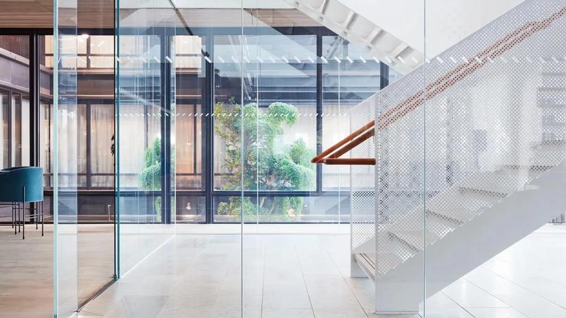 Modern interior with glass walls, a white perforated staircase, and greenery visible through the windows.