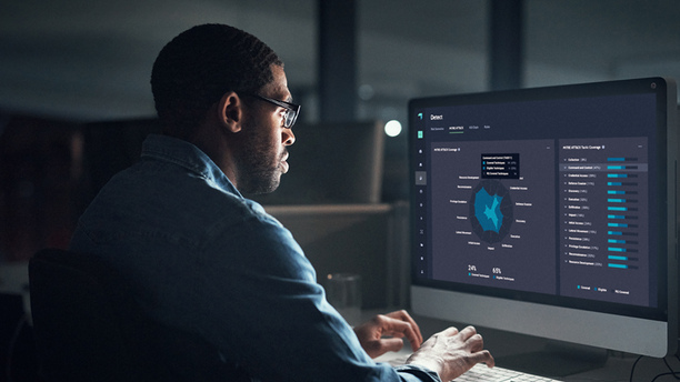 A person wearing glasses sits at a desk in a dimly lit office, looking at a large computer monitor displaying data visualizations and analytics charts on a dark-themed dashboard interface.
