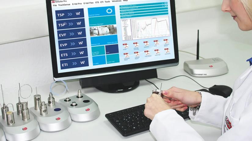 A person in a lab coat working with sensors at a desk, with a computer screen displaying data and graphs.