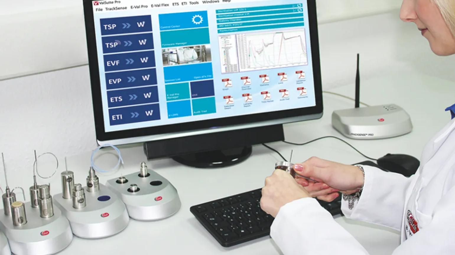 A person in a lab coat working with sensors at a desk, with a computer screen displaying data and graphs.