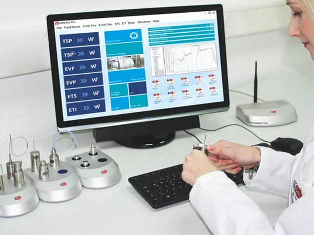 A person in a lab coat working with sensors at a desk, with a computer screen displaying data and graphs.
