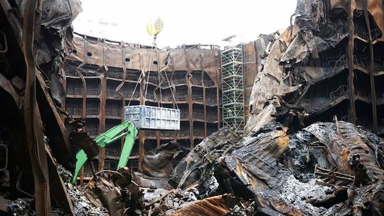 A large industrial site with extensive fire damage, showing twisted metal structures and debris. A green crane is lifting a metal container amidst the wreckage, with scaffolding visible in the background.
