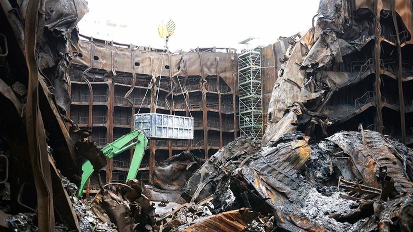 A large industrial site with extensive fire damage, showing twisted metal structures and debris. A green crane is lifting a metal container amidst the wreckage, with scaffolding visible in the background.