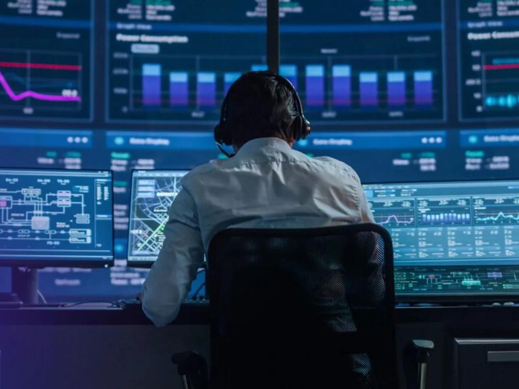 A person wearing headphones sits at a control desk with multiple monitors displaying graphs and charts.