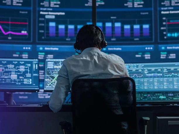 A person wearing headphones sits at a control desk with multiple monitors displaying graphs and charts.