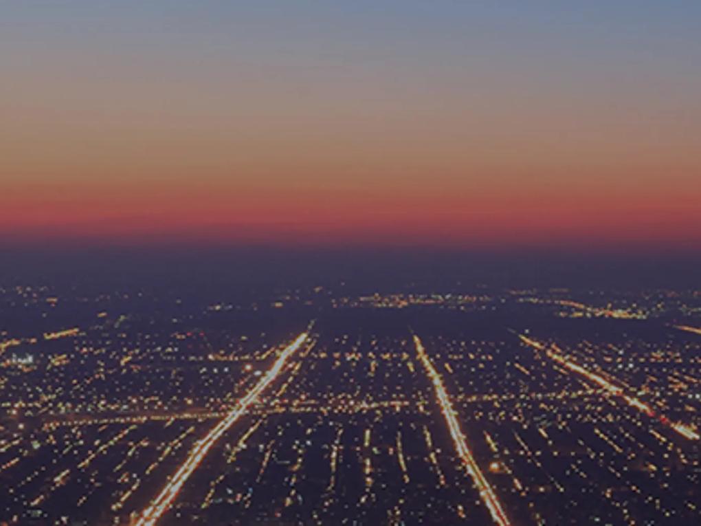 Aerial view of a city at dusk with a grid of illuminated streets and a gradient sky transitioning from orange to blue.