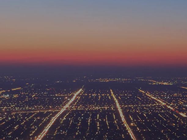 Aerial view of a city at dusk with a grid of illuminated streets and a gradient sky transitioning from orange to blue.