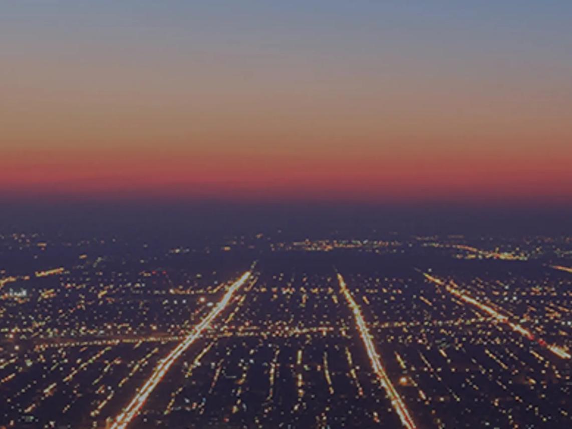 Aerial view of a city at dusk with a grid of illuminated streets and a gradient sky transitioning from orange to blue.