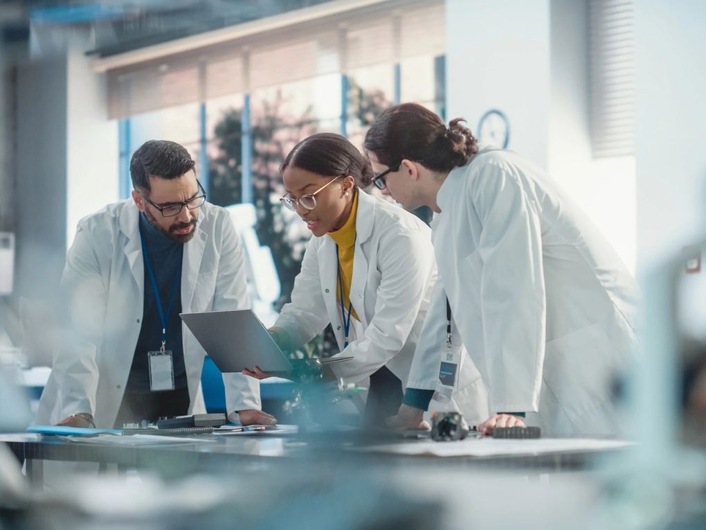 Three scientists in lab coats working together, looking at data on a laptop in a modern research lab.
