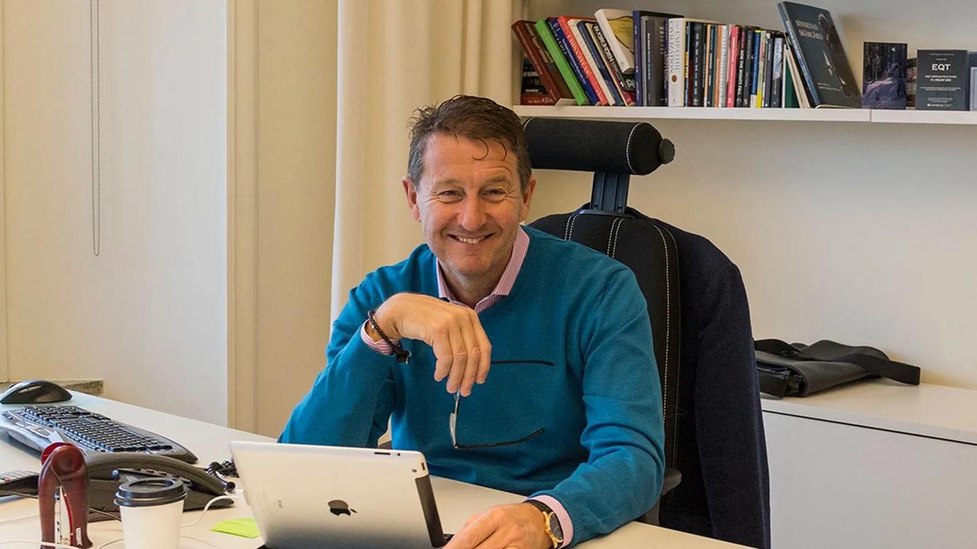 Conni Jonsson sitting at a desk, smiling, with a tablet in front of them. The background features a bookshelf filled with books.