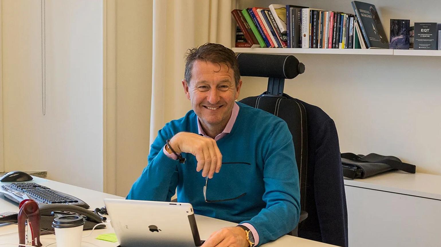 Conni Jonsson sitting at a desk, smiling, with a tablet in front of them. The background features a bookshelf filled with books.
