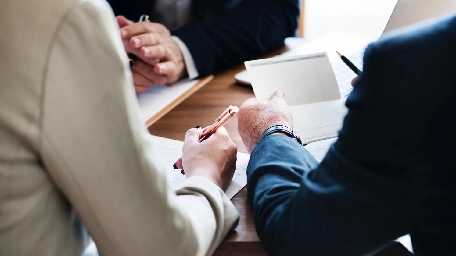 Three people in business attire sitting at a table, discussing documents and taking notes.
