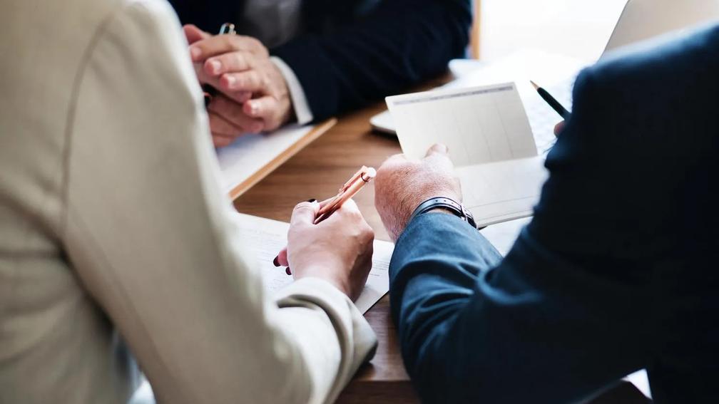 Three people in business attire sitting at a table, discussing documents and taking notes.