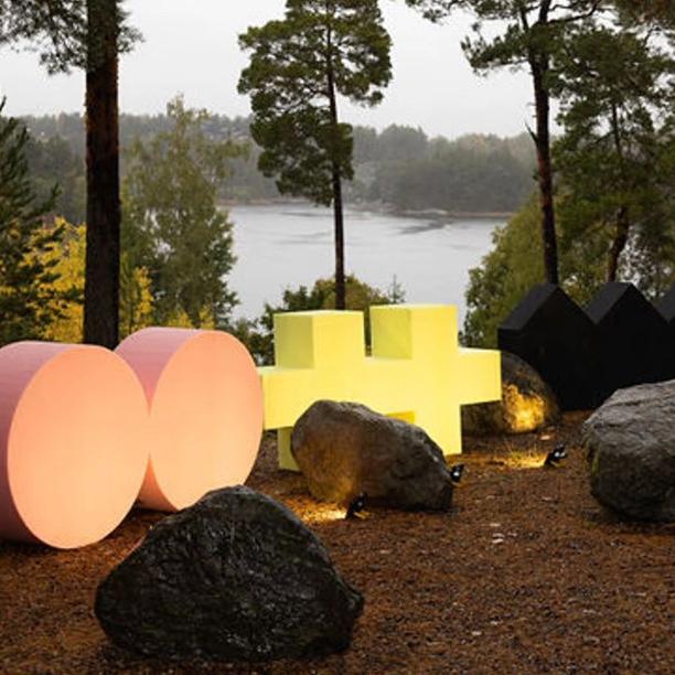Large, colorful geometric sculptures placed among rocks and trees by a lakeside in a forest setting