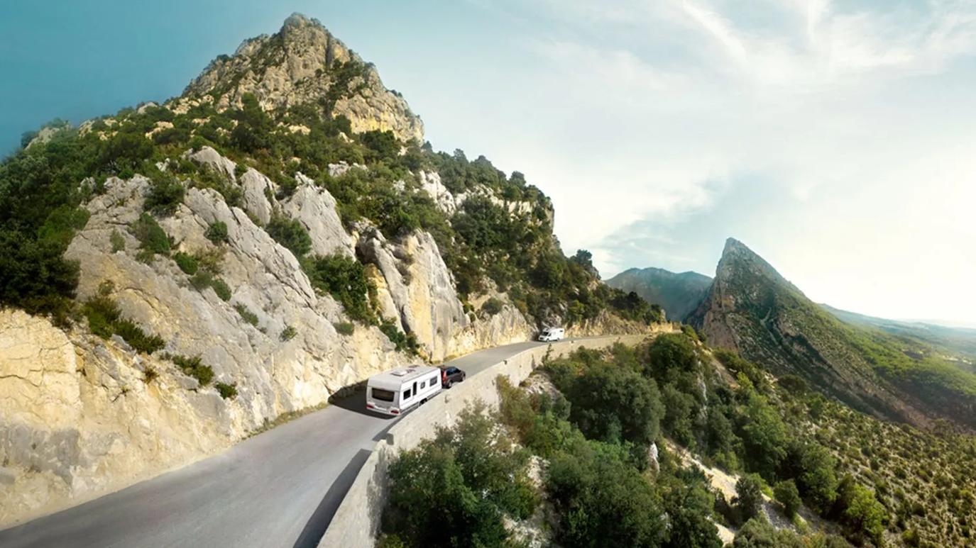 A scenic mountain road with a car towing a caravan, surrounded by rocky cliffs and lush greenery under a clear sky.
