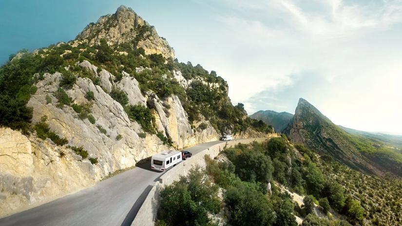A scenic mountain road with a car towing a caravan, surrounded by rocky cliffs and lush greenery under a clear sky.