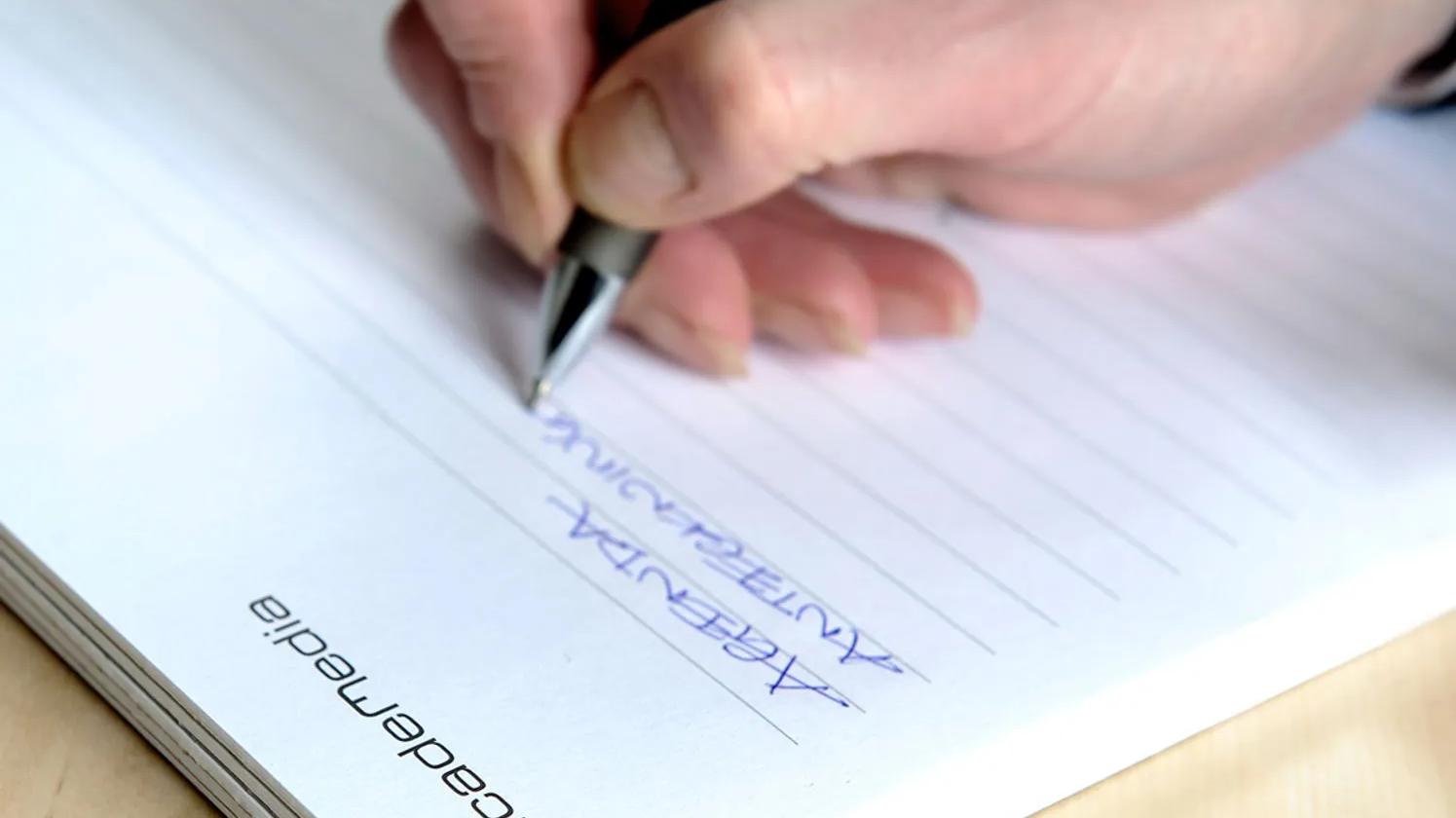 A hand writing with a pen on a lined notepad labeled "academedia," with the word "ARGENTINA" written in blue ink.