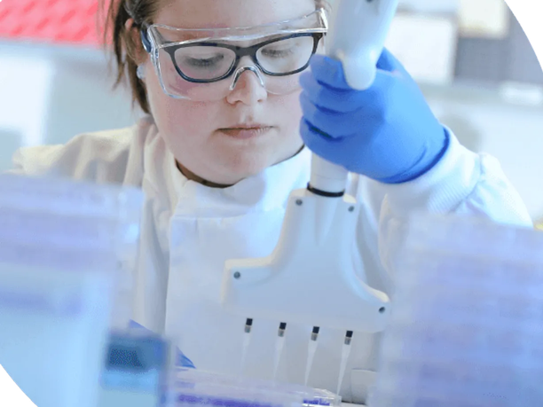 A scientist wearing safety goggles and gloves uses a pipette in a laboratory setting, surrounded by test tubes and lab equipment.