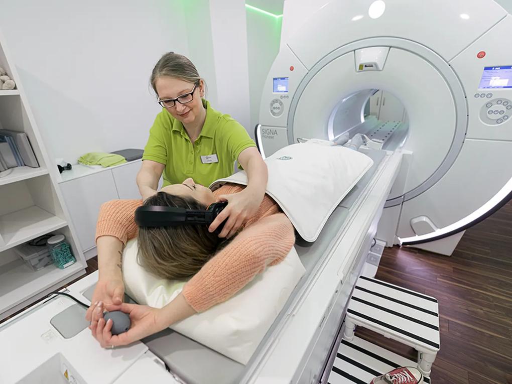 A patient lying on an MRI machine table, wearing headphones, assisted by a technician in a green uniform.