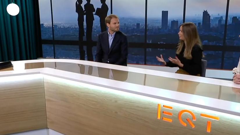 Two people sitting at a modern news desk with "EQT" logo, engaged in conversation, with a city skyline and silhouettes in the background.