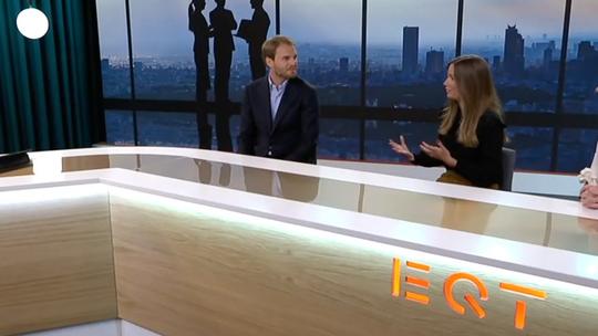 Two people sitting at a modern news desk with "EQT" logo, engaged in conversation, with a city skyline and silhouettes in the background.