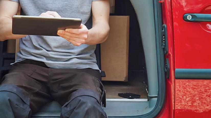 A person sitting in a red car using a tablet, with boxes in the background.