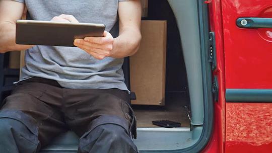 A person sitting in a red car using a tablet, with boxes in the background.