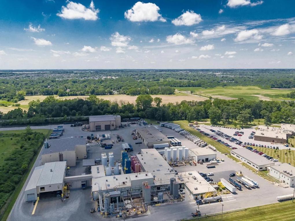 Aerial view of an industrial facility with multiple buildings, storage tanks, and parked vehicles.