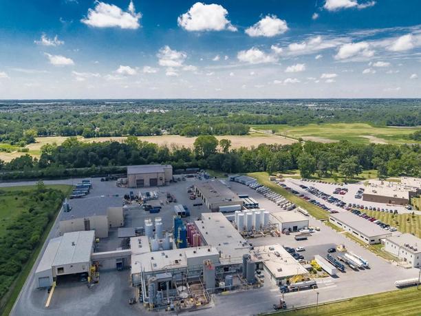 Aerial view of an industrial facility with multiple buildings, storage tanks, and parked vehicles.