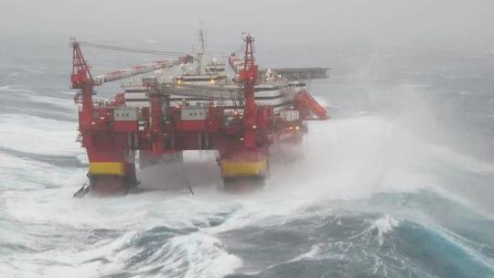 A large offshore oil platform in rough seas, with waves crashing against its structure, highlighting the challenging conditions of ocean-based operations.